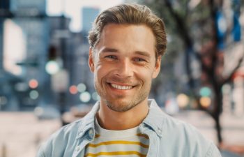 A young man with light brown hair and a beard smiles outdoors, wearing a light blue shirt over a yellow and white striped t-shirt. City buildings and trees appear blurred in the background.