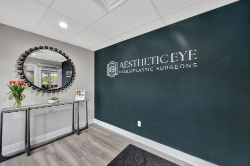A modern reception area featuring a round mirror above a slim console table and a dark wall with the text “Aesthetic Eye Oculoplastic Surgeons.”